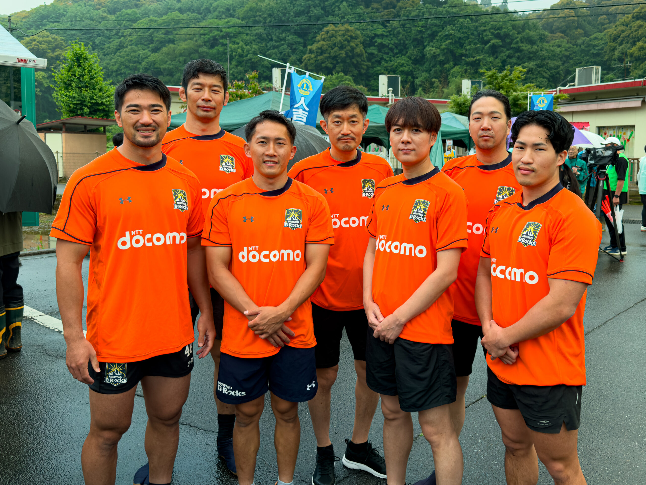 緑の山を背景に、ラグビー選手たちが肩を寄せて立つグループショット