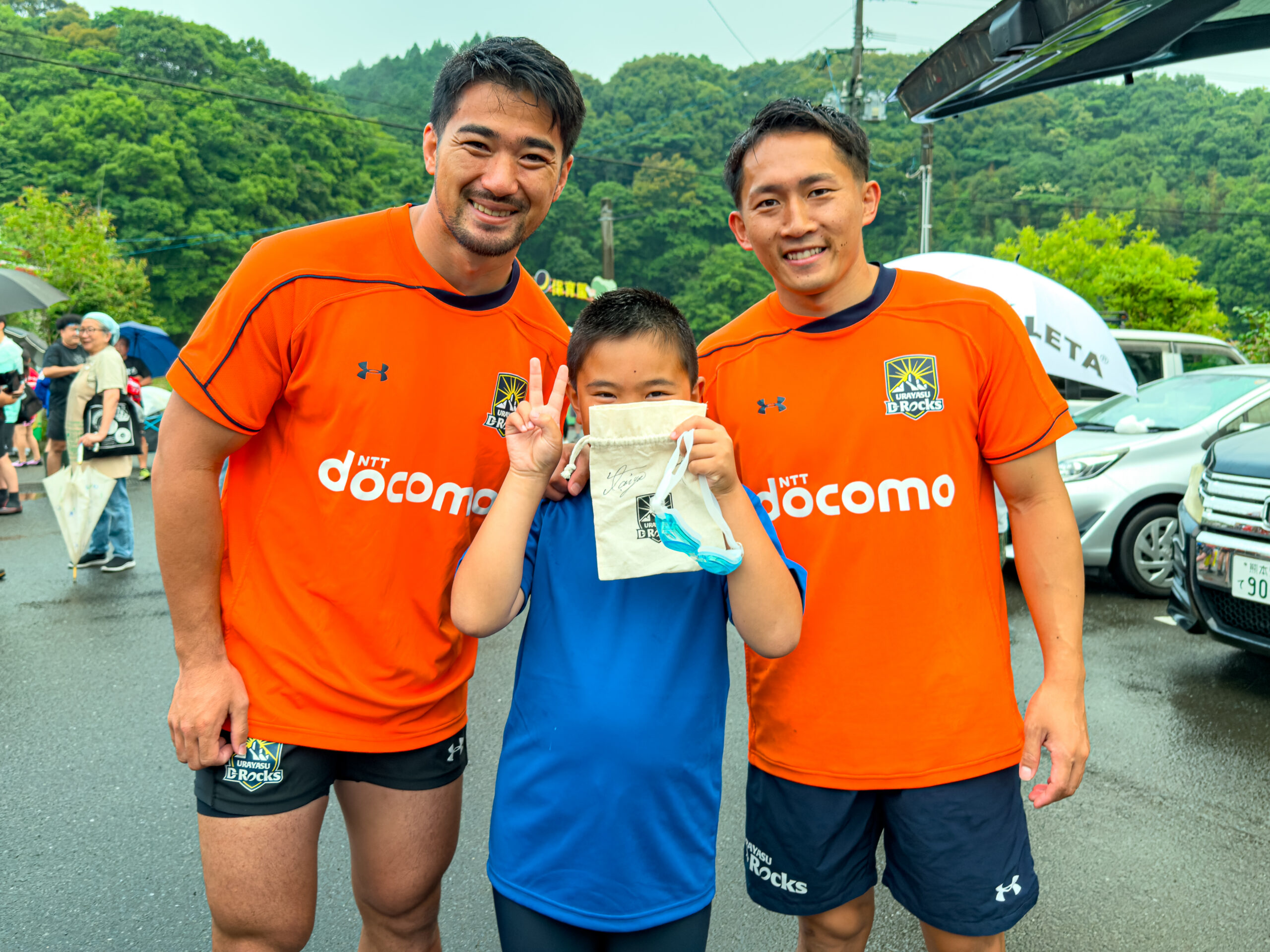 ファンサービス、オレンジのジャージの選手2人と青いシャツの子どもがピースで記念撮影