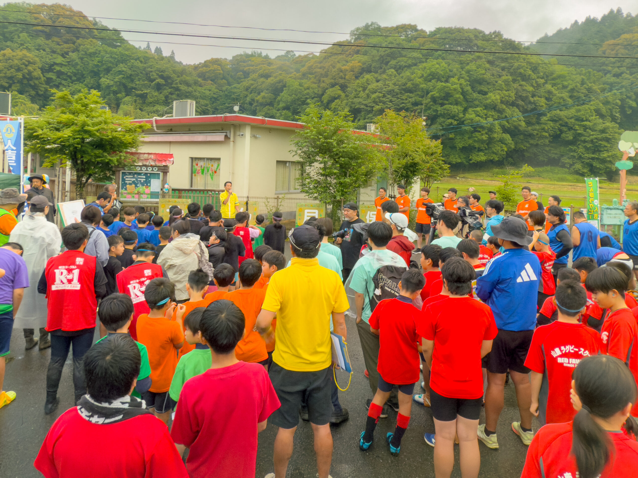 田んぼラグビー大会の開会式、濡れた路面の会場で参加者が集まりスタッフの説明を聞く
