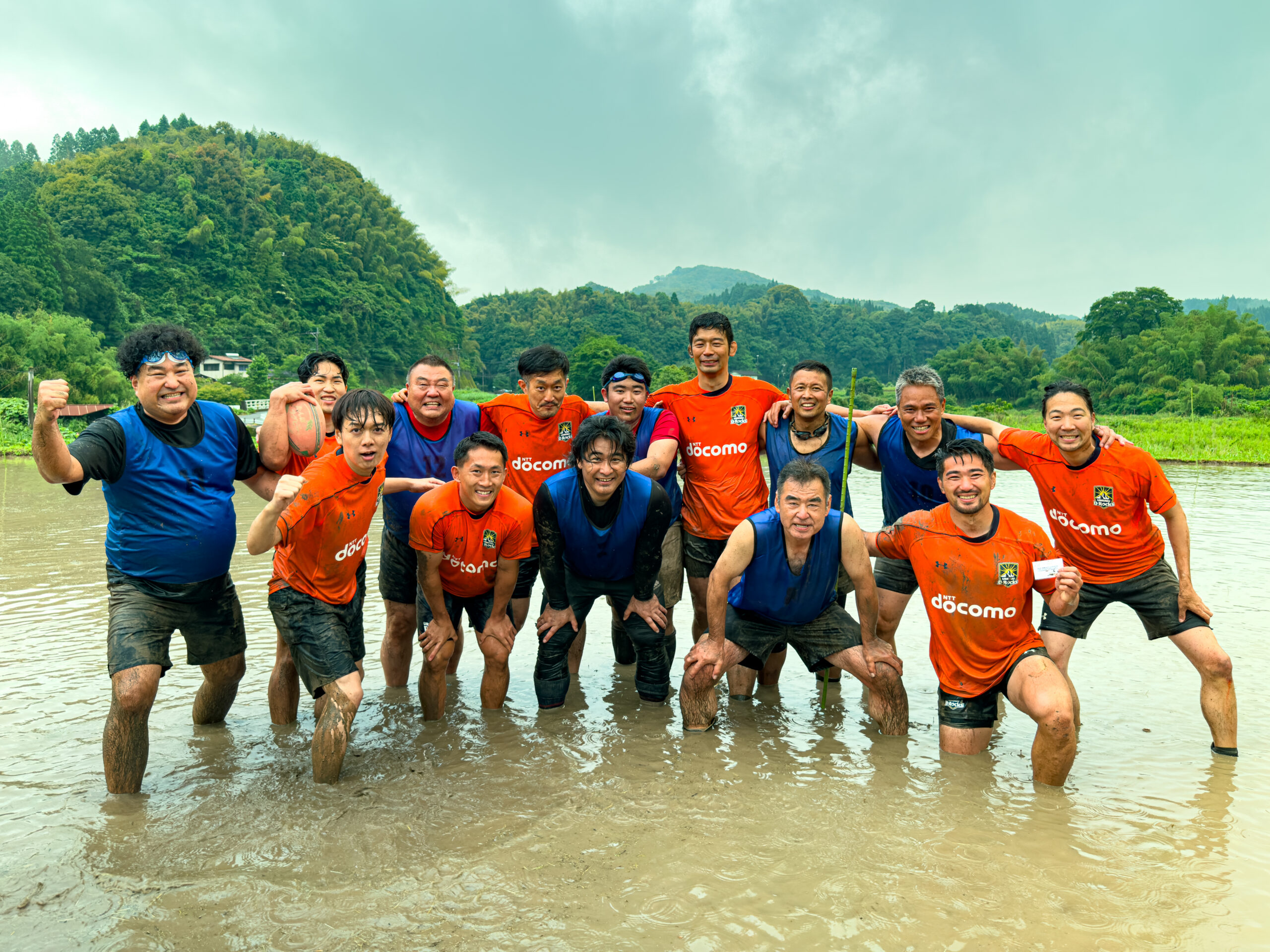田んぼラグビーの選手一同が泥水の田んぼでガッツポーズの集合写真