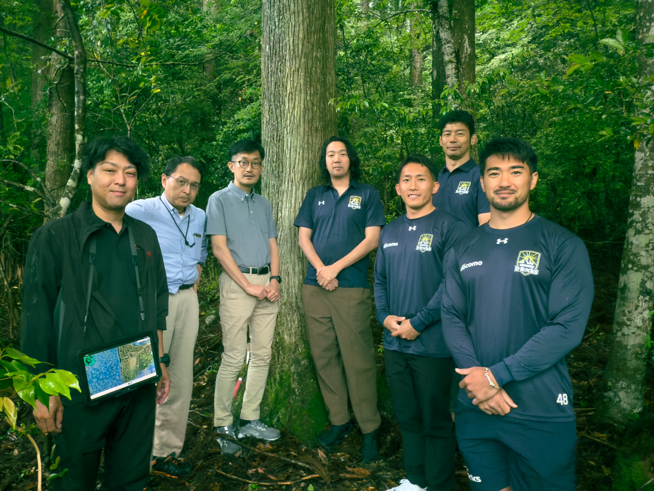 緑豊かな森の中で大木の前に立つメンバー7人の集合写真