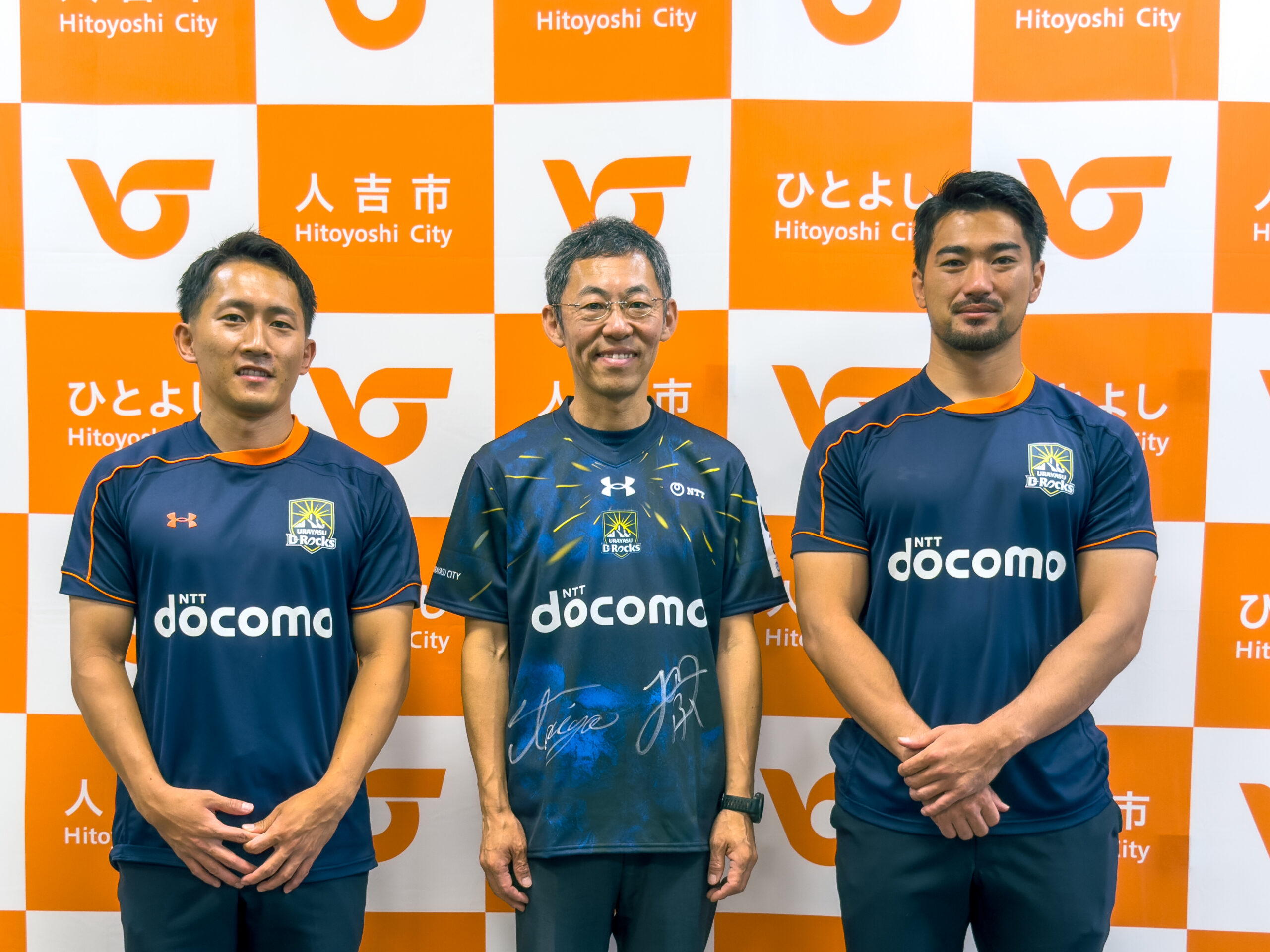 人吉市のパネル前で、スポーツチームのユニフォームを着た3人の集合写真